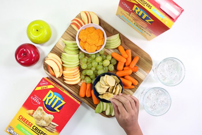 Child reaching for a Ritz Cheese Crisper on a board of snacks,fruits and veggies