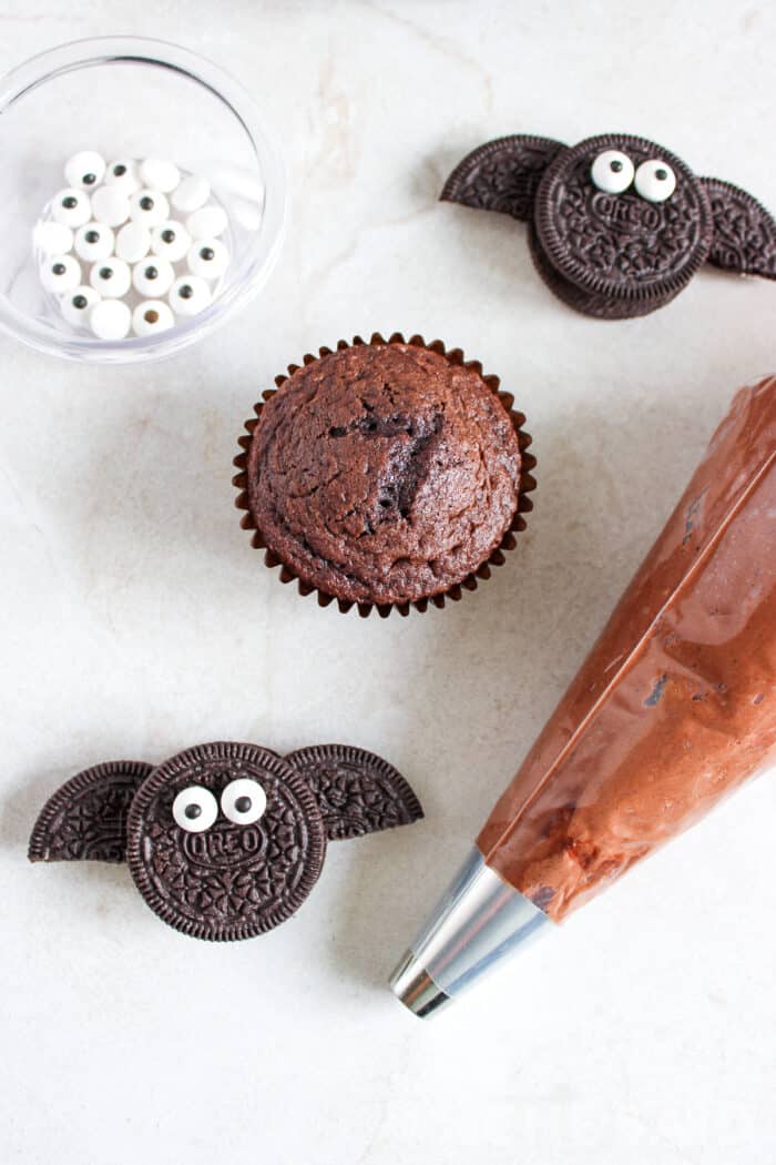 piping bag of chocolate butter cream frosting, a naked chocolate cupcake and an oreo bat ready for assembling